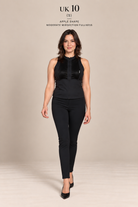 A woman modeling a black sleeveless top and black pants against a plain beige studio background. Text at the top reads 'UK 10 (S)' and 'APPLE SHAPE MODERATE MOSSECTION FULLNESS'.